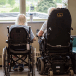 Two residents sitting in mobility devices looking out the window of the GPL.