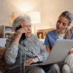 Older woman on the phone with a laptop on her left being supported by a young woman. 