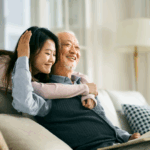 Young woman hugging an older man sitting down on a couch. 